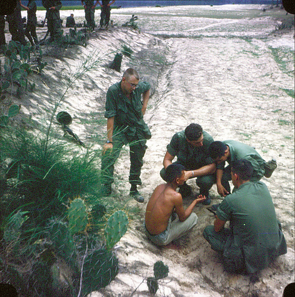 On The Beach
My failing memory suggests that this was before we went into the mountains [and carnage] west of Tam Ky, so we were still relatively 'unbloodied' except for a few scrapes. Maybe October, 67? For a day or two, I think we were kind of pinned in with the sea at our back, on a white sand beach that was very easily defended. Choppers brought is food and mail and flew in from the sea instead of  over land for the 2-3 days were were there. Even night was fairly safe since we had nearly a full moon, clear skies and anything that moved was pretty easily spotted in the white sand. Does any of that sound familiar? 
(Bert watches intently as a "local" is giving information.  Note his typically discarded helmet appears in the rear; could never get one that fit. ) 
